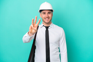 Young architect caucasian man with helmet and holding blueprints isolated on blue background happy and counting three with fingers