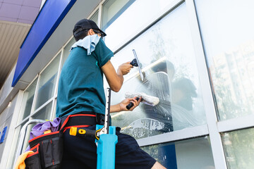 A young male worker of a professional cleaning service in overalls washes the glass of the windows...