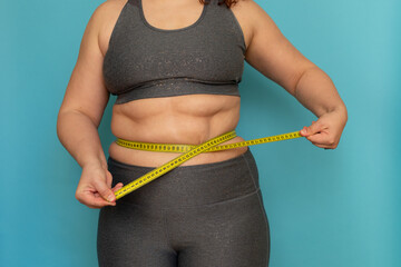 Cropped photo of fat plump overweight woman standing in grey sports bra and leggings, showing excess naked belly, measuring waist, holding tape on blue background. Dieting, unhealthy food, obesity.