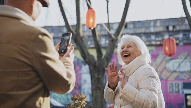 Elderly couple takes photos smiling on a smartphone on a modern street. Senior couple learning new technologies and practicing photography on a phone. High quality 4k footage