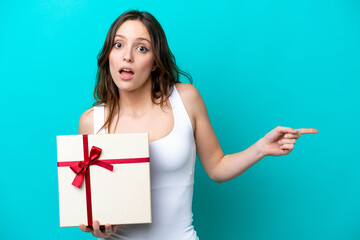 Young caucasian woman holding a gift isolated on blue background surprised and pointing finger to the side