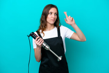 Young caucasian cooker woman using hand blender isolated on blue background with fingers crossing and wishing the best