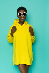 Smiling young black woman in sunglasses and vibrant yellow oversized hooded sweatshirt.