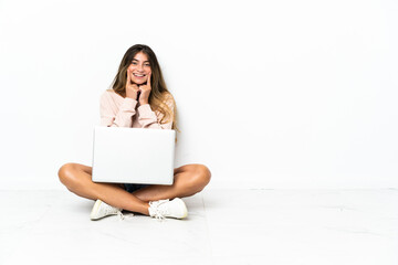 Young woman with a laptop sitting on the floor isolated on white background smiling with a happy and pleasant expression