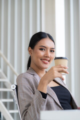 Portrait of beautiful female employee sitting happily smiling towards the camera holding a ready-to-drink paper coffee cup ready for the start of daily office work