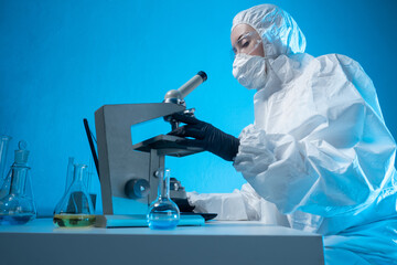 Biologist at work. Chemist next to microscope. Woman in hazmat suit. Biological lab worker. Biologist sits at table with test tubes. Laboratory assistant on blue background. Research in microbiology