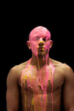 Portrait of young muscular man covered with thick pink and yellow paint isolated over black background
