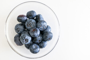 Top view of many juicy blueberries in a glass bowl on the left side of white table background. A sweet, delicious and healthy fruit delight