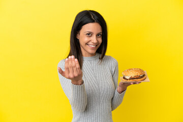 Young caucasian woman holding a burger isolated on yellow background inviting to come with hand. Happy that you came