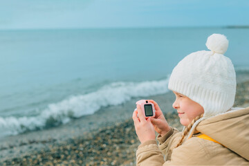 a little girl squinting photographs the autumn seascape on a childrens pink camera. the child is...