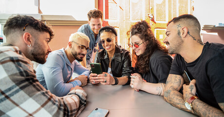 Group of young fashionable friends sitting outdoors and looking at mobile phone, watching social media. Smiling students having fun together.