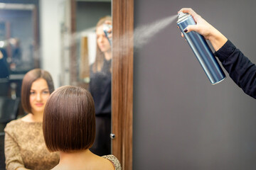 A hairdresser is using hair spray to fix the short hairstyle of the young brunette woman sitting in...