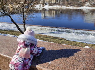 A little girl looks at the river after the spring ice drift. Photo of a child from the back in sunny weather. Plain air and landscapes in spring.