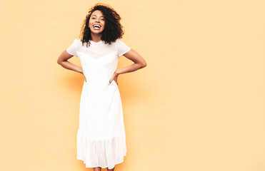 Portrait of beautiful black woman with afro curls hairstyle. Smiling model dressed in white summer dress. Sexy carefree female posing near yellow wall in studio. Tanned and cheerful. Isolated