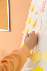 Woman writing on sticky note attached to flipchart near beige wall, closeup - obrazy, fototapety, plakaty