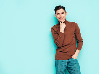 Portrait of handsome smiling model. Sexy stylish man dressed in turtleneck sweater and jeans. Fashion hipster male posing on grey background in studio. Isolated. Touching his chin