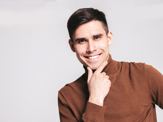 Portrait of handsome smiling model. Sexy stylish man dressed in turtleneck sweater and jeans. Fashion hipster male posing on grey background in studio. Isolated. Touching his chin