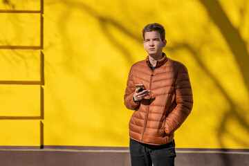 a young man in a brown jacket stands on the street  against a yellow wall. Young man holding phone...