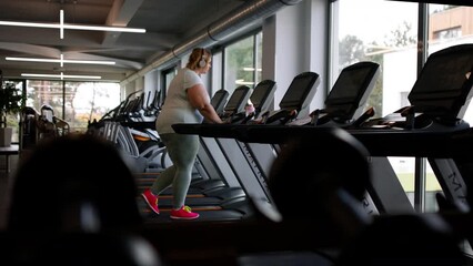 Overweight woman with headphones exercising on treadmill in gym - Powered by Adobe