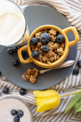 granola oatmeal with blueberries in yellow bowl on blue plate close-up, glass of milk, yogurt and yellow tulips on striped tablecloth,  healthy colorful breakfast