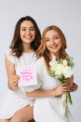 Young woman and her mother with flowers and greeting card for International Women's Day on light background