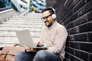 A happy man with headphones sits on the stairs outside and using laptop.
