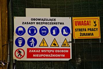 Tablica z obowiązującymi na budowie zasadami bezpieczeństwa w formie piktogramów . Tabliczka : " Uwaga strefa pracy żurawia " .