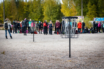 a group of people watching the disc golf demo in the park. two disc golf baskets in front