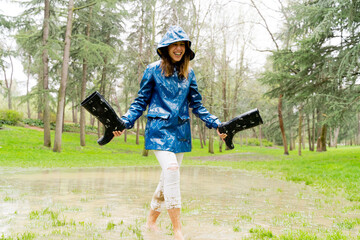front view of woman walking barefoot on pond holding galoshes. Horizontal panoramic view of woman splashing with mud on rainy pond. Nature and people backgrounds.