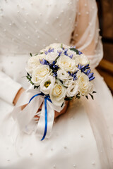 Wedding bouquet of white roses and blue flowers with a blue ribbon in the hands of the bride on the background of the wedding dress, close-up, details at the wedding