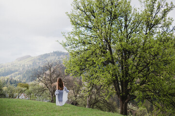 Young pretty emotional caucasian woman with long hair wearing natural makeup in romantic casual clothes walking among spring mountains near the beautiful fresh blooming tree in the countryside.