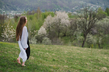 Naklejka na ściany i meble Young pretty emotional caucasian woman with long hair wearing natural makeup in romantic casual clothes walking among spring mountains near the beautiful fresh blooming tree in the countryside.