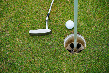 Obraz premium Top view of a golf ball with putter on green course at hole. Golf ball and golf club on green in the evening golf course with sunshine.