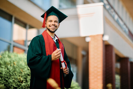 Happy university student in graduation gown holding his diploma and looking at camera.