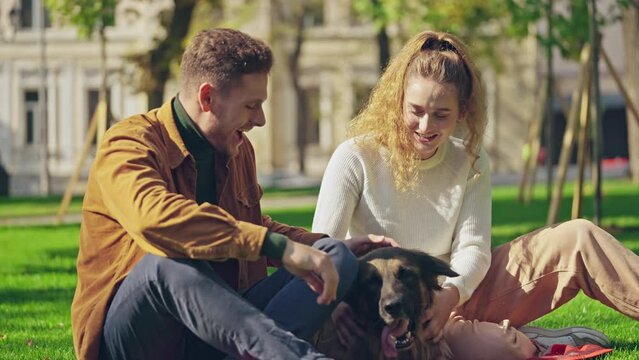 Young man and woman sitting in park, hugging and petting german shepherd dog