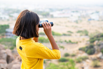 Girl kid watching far destinations by using binocular at hill top during summer camp - concept of exploration, expedition and vacation.