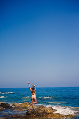 Beautiful young woman in a swimsuit on a rocky beach on a sunny day against the backdrop of waves. Vacation in the summer season. Selective focus