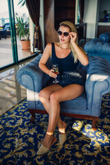 Beautiful young woman sits on a soft armchair in the hotel lobby with a glass of champagne on vacation. She is wearing sunglasses and a smile on her face. Vacation concept
