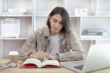 Young male student reading a book in a private office, Long distance education, Textbooks from books, Find out more, In the office there is a computer or laptop placed on the table, Study.