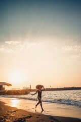 Young beautiful woman in a black swimsuit and hat with glasses walks along the beach at sunset. The concept of sea recreation. Selective focus