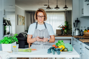 Woman using phone and looking for recipe online for cooking dinner in kitchen at home. Watching tutorial video while preparing healthy dish with fish and vegetables. Balanced dieting. Selective focus.