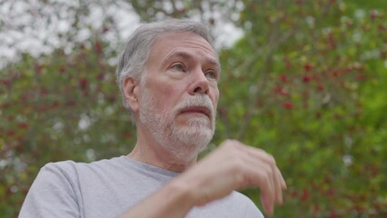 A senior man standing outdoors starts sneezing and rubbing his eyes as his allergies react to all the seasonal plant pollen in the air.