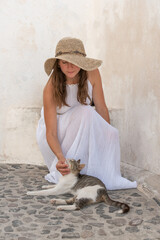Young beautiful woman in white long dress and sun hat touching a street cat in Santorini greek...
