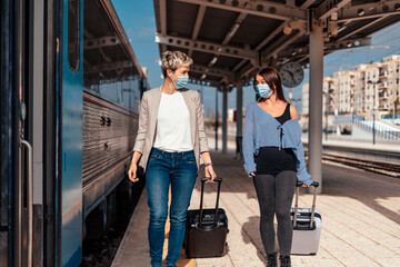 Two happy female friends in protective masks  walking and chatting at train station