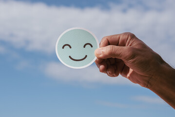 A paper-cut smiling face in a man's hands on a beautiful blue sky and white cloud in sunny day with natural light, Positive picture or happy  life or World mental health day and eco friendly concept.