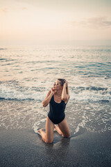 Attractive young woman in sunglasses kneels in the sea at sunset light in a black swimsuit. Selective focus