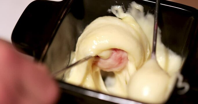 two hands with forks dip the cherry tomatoes into the melted fundu cheese in a black plate and pull the cheese up. view from above. close-up