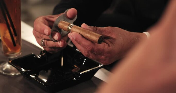 male hands with a gold ring on their finger are cutting a cigar with a guillotine of scissors, on a table near a cocktail over an ashtray. close-up