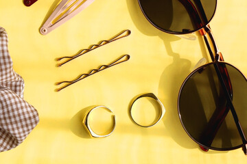 Retro sunglasses, gold rings, various hair accessories and make up products on vibrant yellow background. Flat lay.