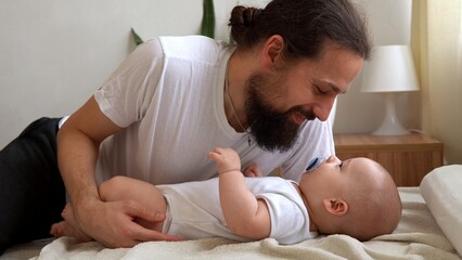 Authentic Young Bearded Man Holding Newborn Baby. Dad And Child Son On Bed. Close-up Portrait of Smiling Family With Infant On Hands. Happy Marriage Couple On Background. Childhood, Parenthood Concept